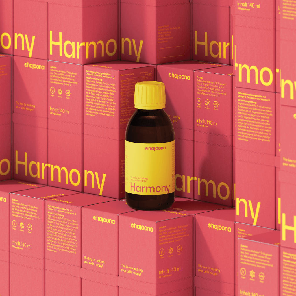 Eine braune Flasche hajoona Harmony mit gelbem Deckel und gelbem Etikett steht vor einem Hintergrund aus gestapelten korallenfarbenen Verpackungen mit gelber Schrift. Das Design hebt die Premium-Qualität und Natürlichkeit des Produkts hervor.