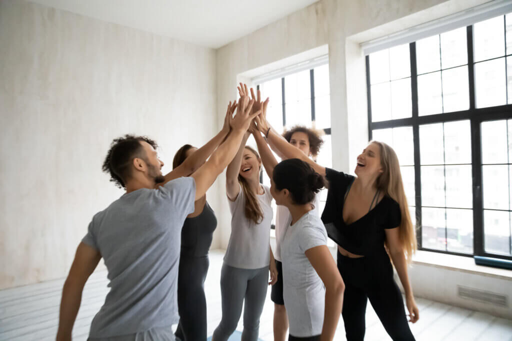 Eine Gruppe von sechs Personen steht in einem hellen Raum mit großen Fenstern. Sie tragen sportliche Kleidung in Grau, Schwarz und Weiß. Alle strecken ihre Hände in die Mitte und berühren sich gegenseitig, während sie lachen. Das Bild zeigt Freude, Teamgeist und Motivation.